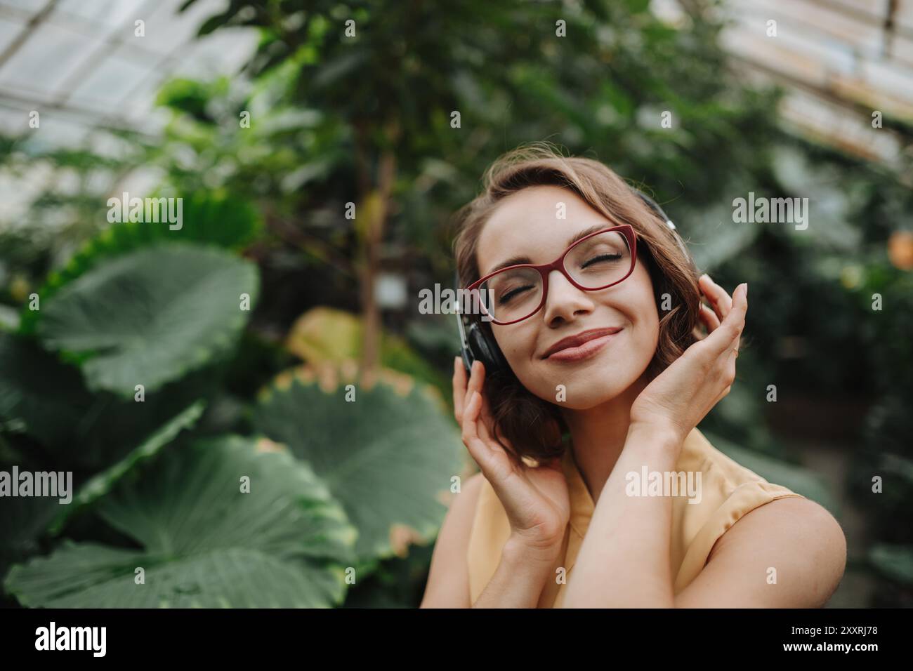 Schöne Frau, die im botanischen Garten steht, mitten in üppig grünem Laub mit Kopfhörern auf dem Kopf, Musik hört. Stockfoto