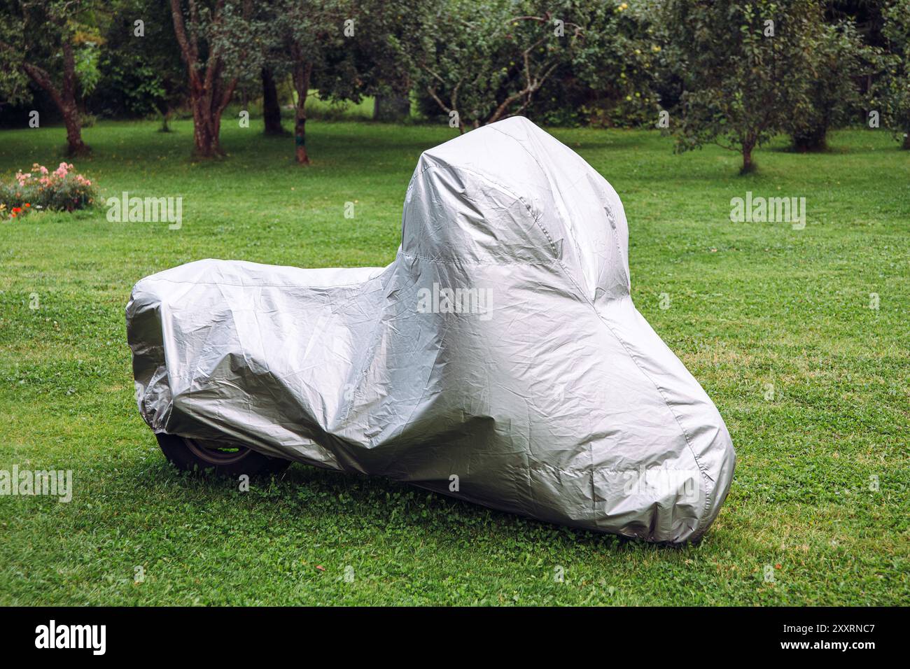Das Motorrad parkt im Garten und ist mit einer grauen wasserdichten Motorradabdeckung bedeckt. Stockfoto