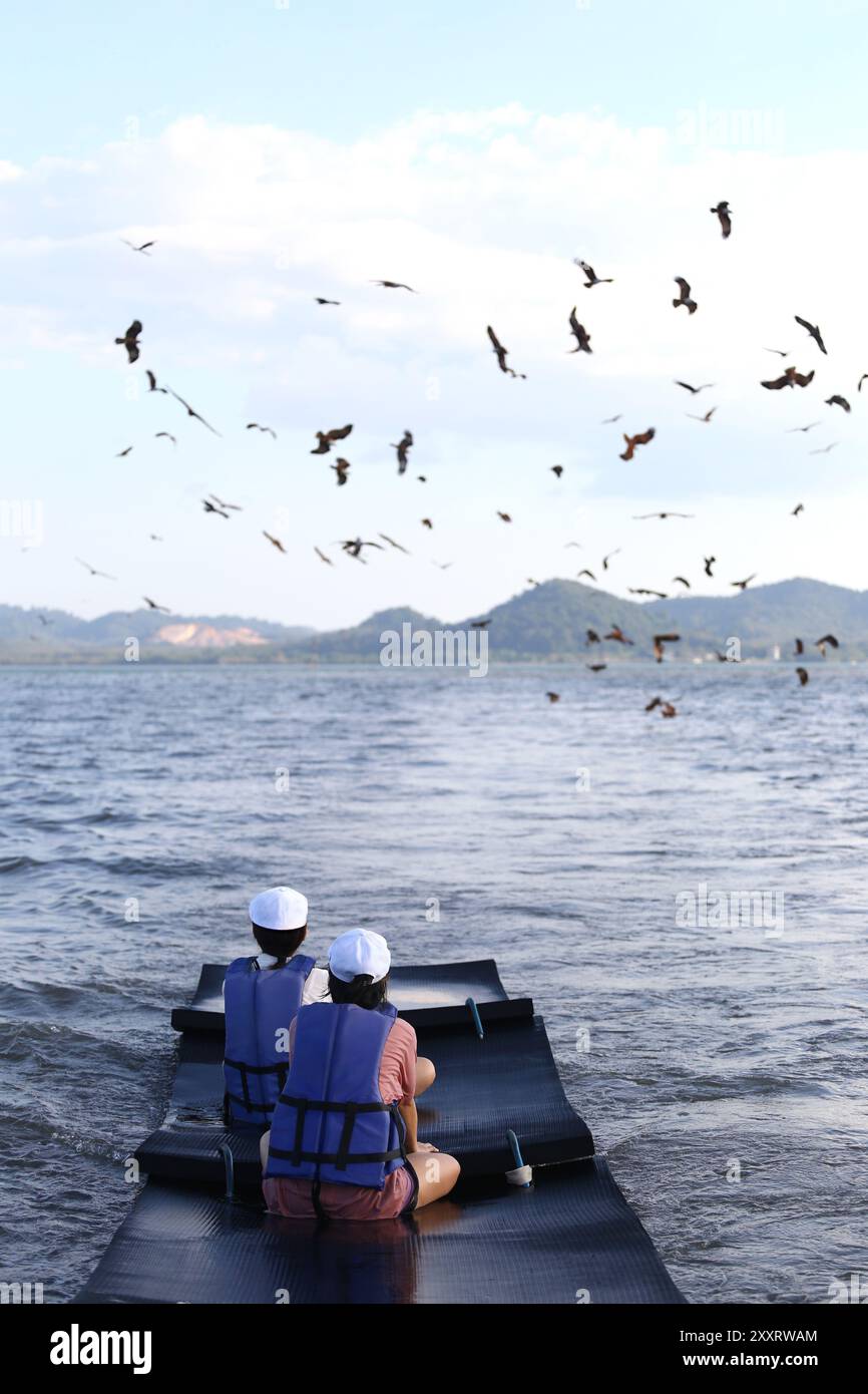 Zwei Touristen setzen eine Schwimmweste auf einem Floß mitten im Meer und sehen, wie viele Haliastur indus in der Provinz Chanthaburi in den Himmel fliegen. Stockfoto