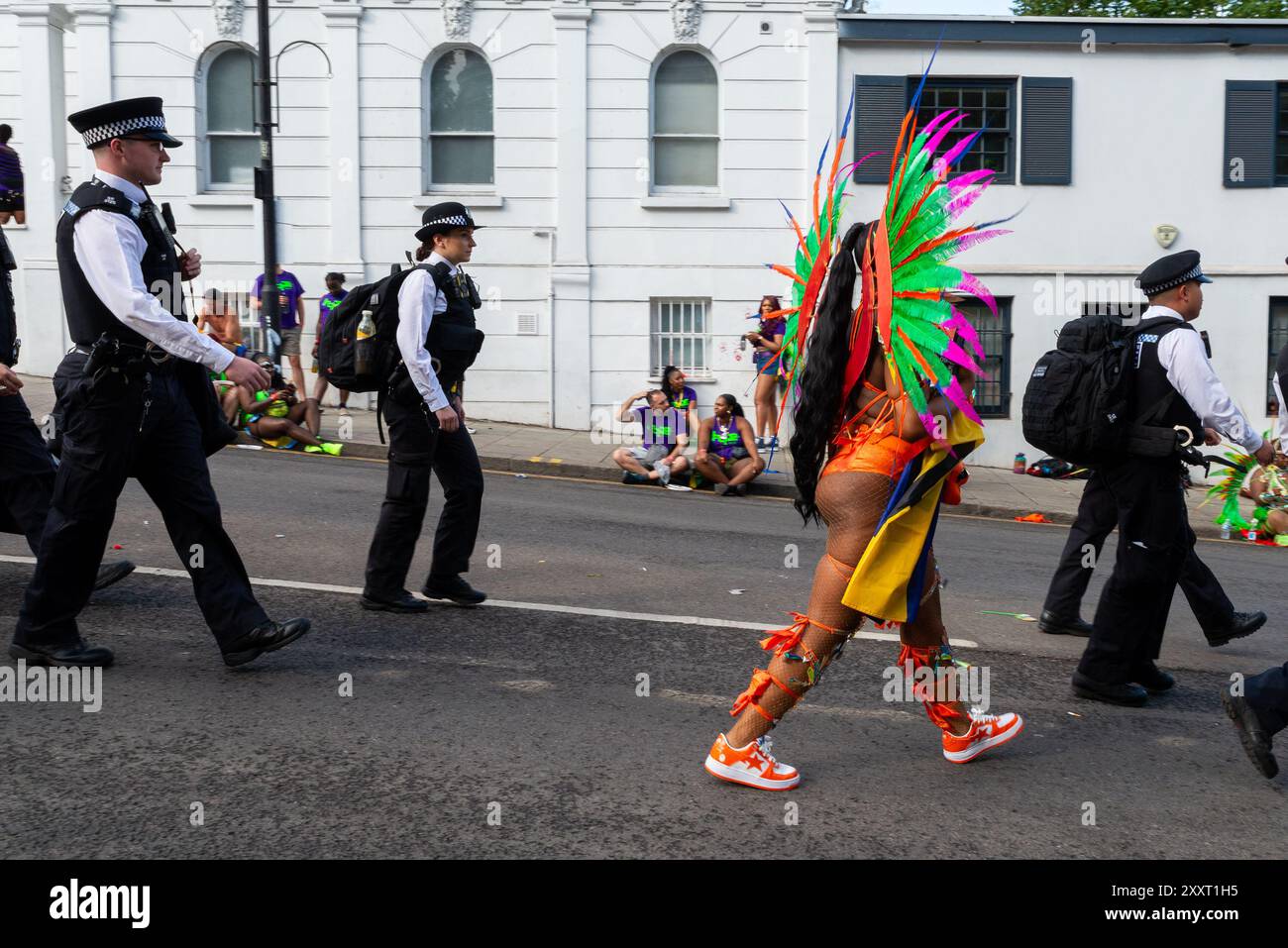 Notting Hill, London, Großbritannien. August 2024. Europas größtes Straßenfest findet später in den Straßen von Notting Hill statt. Exotische Tänzer und Musikgruppen im jamaikanischen Stil ziehen durch die Straßen, während Speisen und Unterhaltung in der Gegend zur Veranstaltung beitragen. Die Grand Parade findet am Montag der Bank als Höhepunkt des dreitägigen Festivals statt, das 1966 begann. Die Teilnehmer versammeln sich in der Nähe und bereiten ihre farbenfrohen Kostüme vor, mit Sicherheitspolizisten und Graffiti-bedeckten Schutzschildern. Die Polizei kommt an Stockfoto