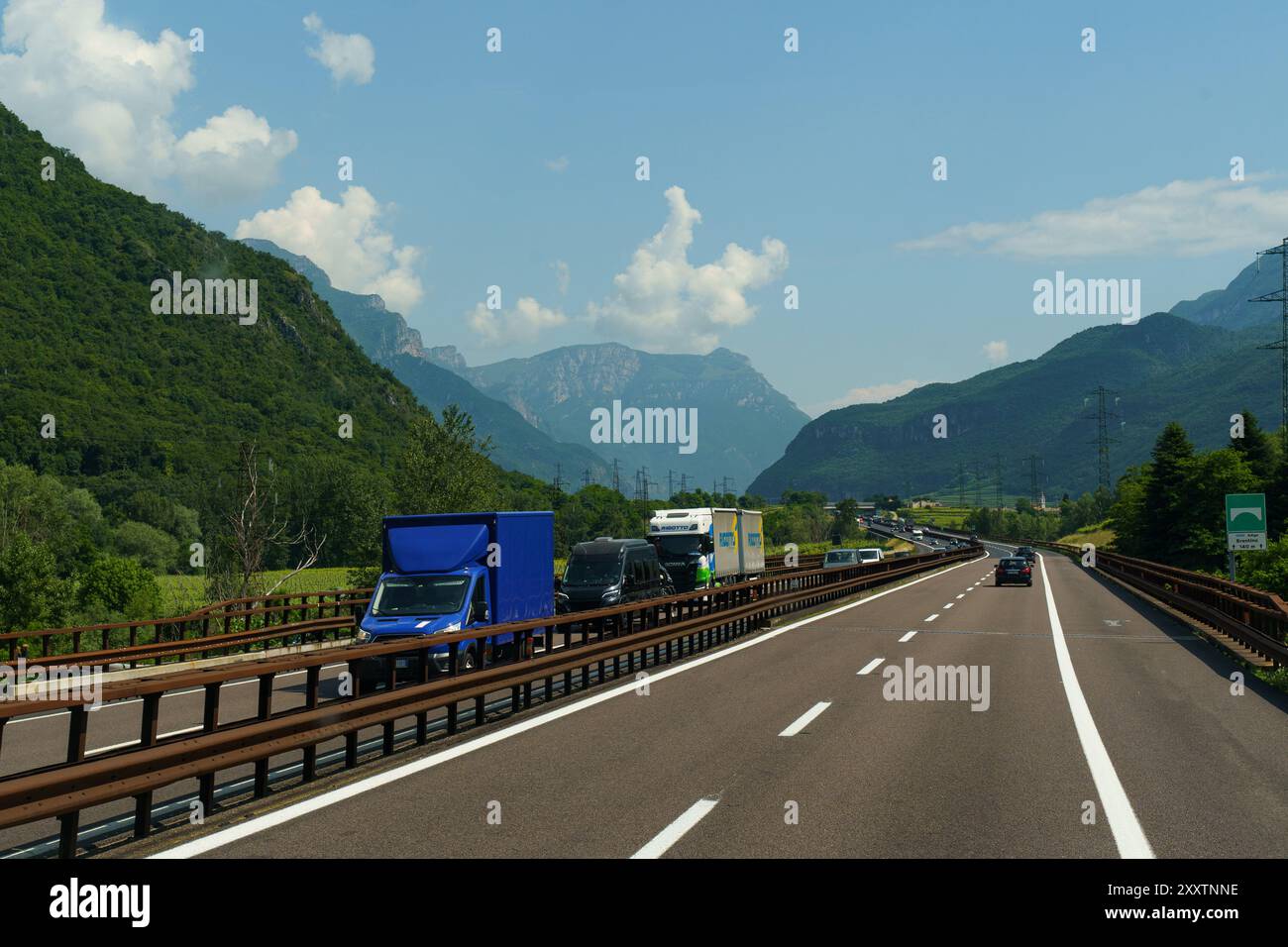 Avio, Italien - 8. Juni 2023: Fahrzeuge fahren entlang einer Autobahn, flankiert von lebhaftem Grün und majestätischen Bergketten unter klarem blauem Himmel. Stockfoto