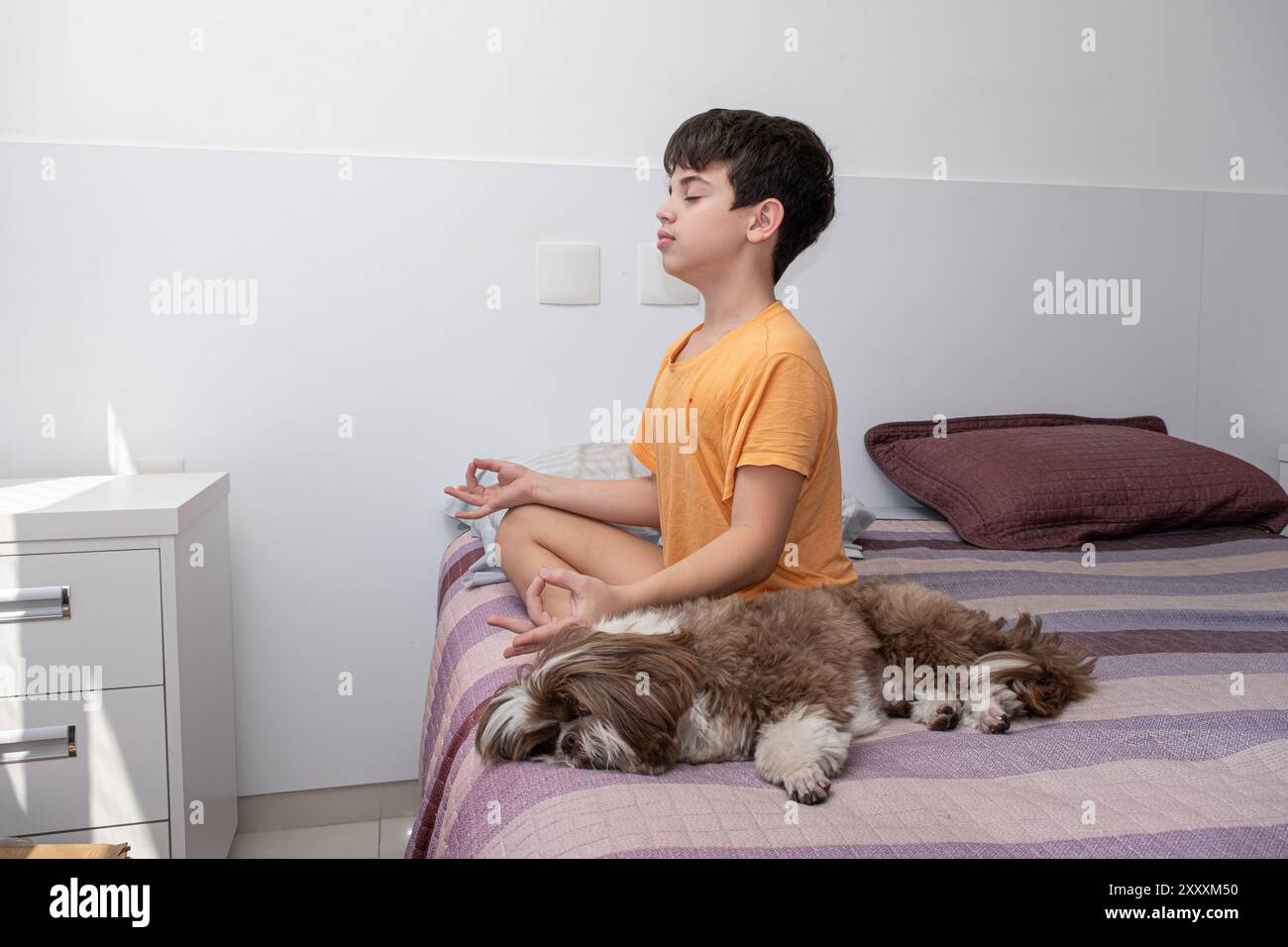 Der 11-jährige brasilianische Junge sitzt auf seinem Bett und übt Yoga neben seinem Hund 1. Stockfoto