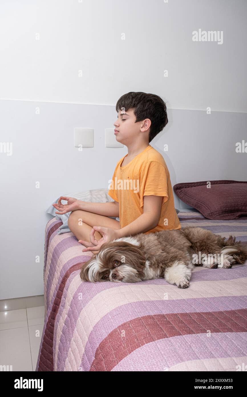 Der 11-jährige brasilianische Junge sitzt auf seinem Bett und übt Yoga neben seinem Hund 3. Stockfoto