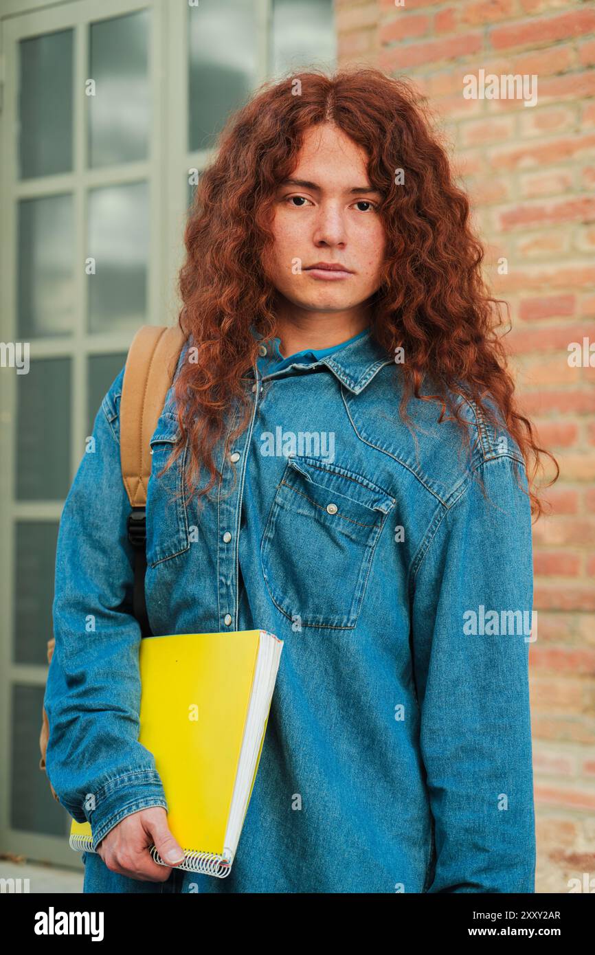 Vertikales Einzelporträt eines ernsthaften Studenten, der auf dem Universitätscampus steht und in die Kamera blickt. Nachdenklicher Teenager, der ein Notizbuch in der Highschool hält. Junge Erwachsene Person, die auf die Akademie starrt. Hochwertige Fotos Stockfoto