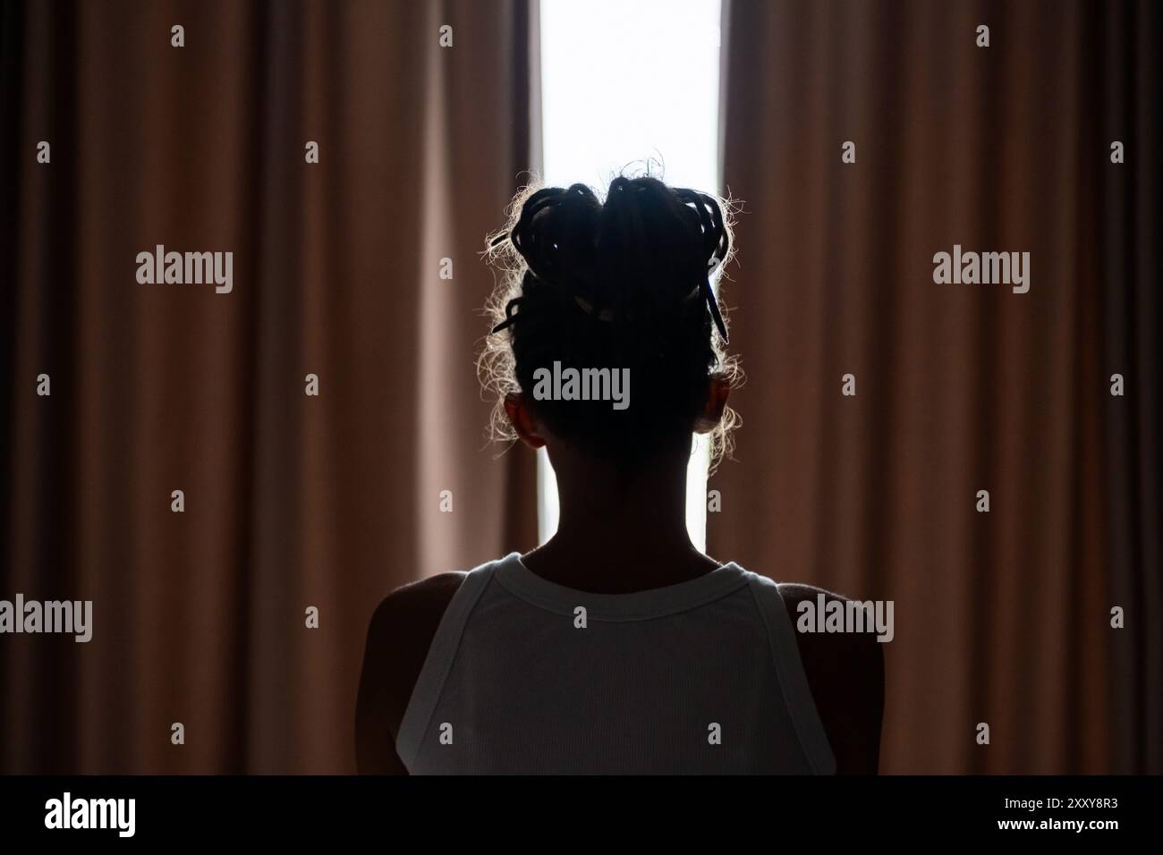 Weibliche Silhouette mit Bun-Frisur, die vor dem Fenster in schwach beleuchtetem Raum steht. Stockfoto