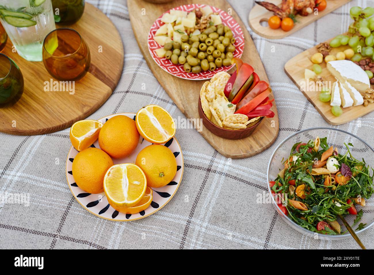 Frisches Obst, Käse, Oliven und ein lebendiger Salat auf einer Picknickdecke mit hölzernen Servierbrettern und Gläsern schaffen eine einladende Atmosphäre im Freien für eine entspannende Mahlzeit Stockfoto