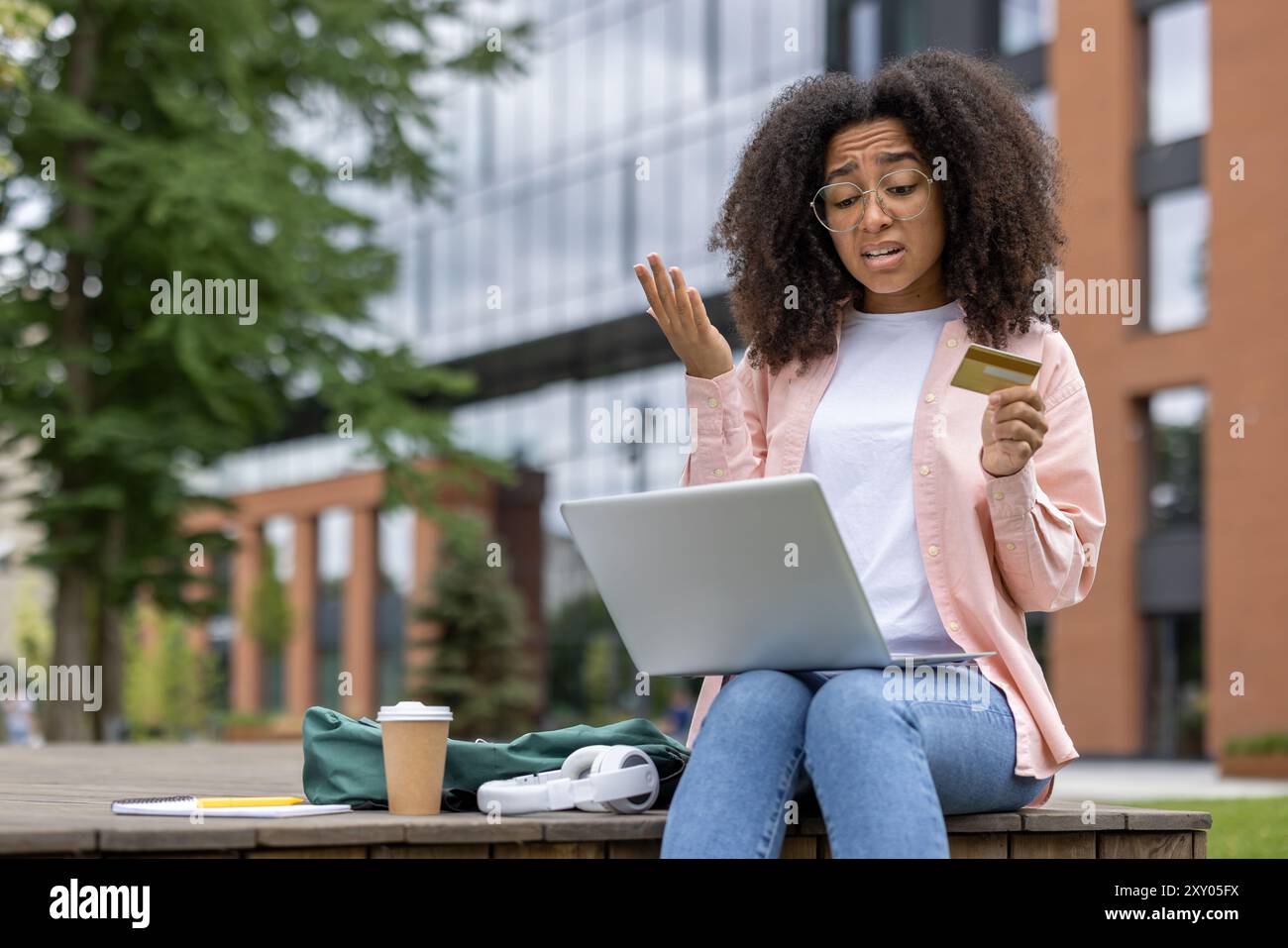 Verwirrte junge Frau mit Laptop und Kreditkarte im Freien. Frustration während der Online-Shopping-Transaktion auf der Campus-Bank zeigen. Bäume und modernes Gebäude im Hintergrund. Stockfoto