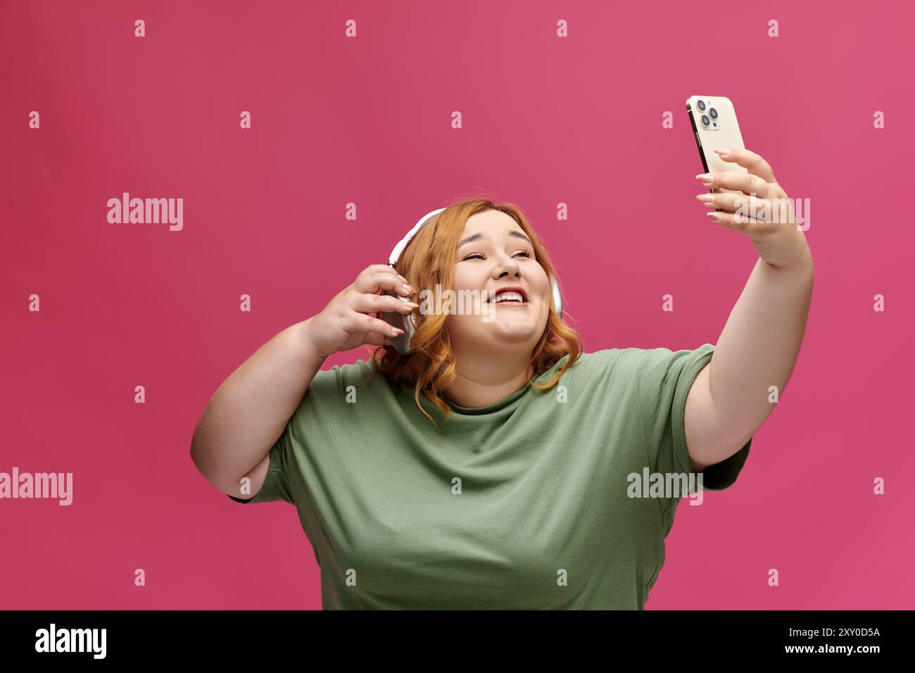 Eine Frau in einem grünen Hemd und Kopfhörern macht ein Selfie mit einem Smartphone, während sie lächelt. Stockfoto