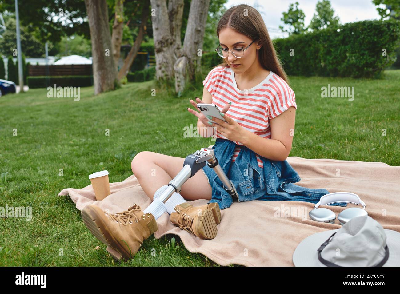 Eine junge Frau mit einer Beinprothese genießt einen sonnigen Tag im Park, während sie ihr Telefon überprüft und sich auf einer Decke entspannt. Stockfoto