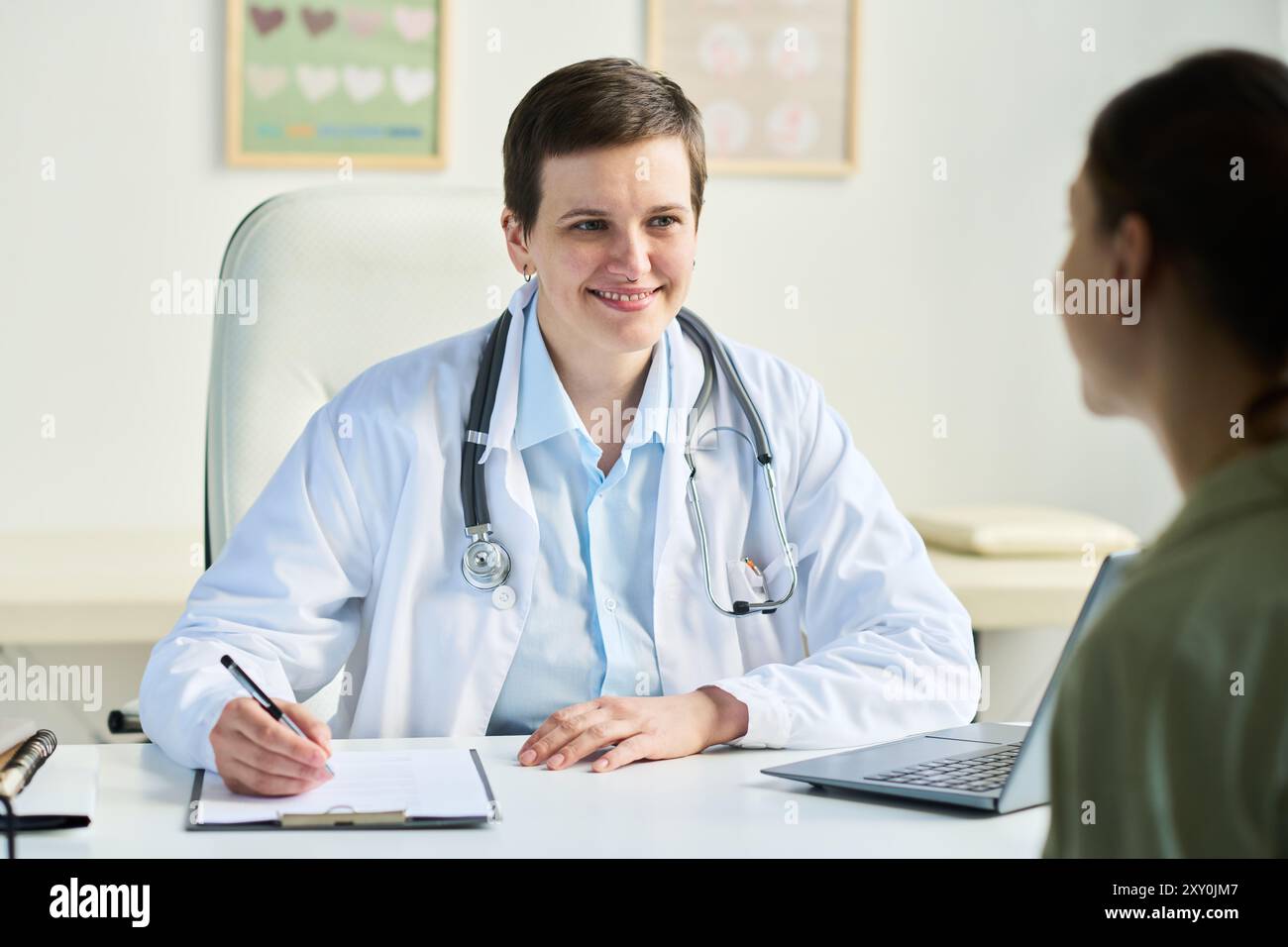 Porträt einer leitenden Ärztin mit weißem Mantel und Stethoskop, die sich in freundlichem Gespräch mit dem Patienten einlässt, während sie Notizen im Büro mit medizinischen Akten im Hintergrund macht Stockfoto