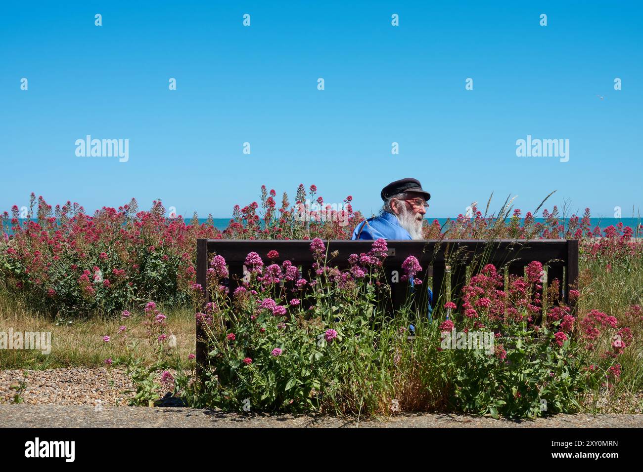 Alter Mann mit Fischerhut, der auf einer Bank am Strand in Aldeburgh, Suffolk, England sitzt. Stockfoto