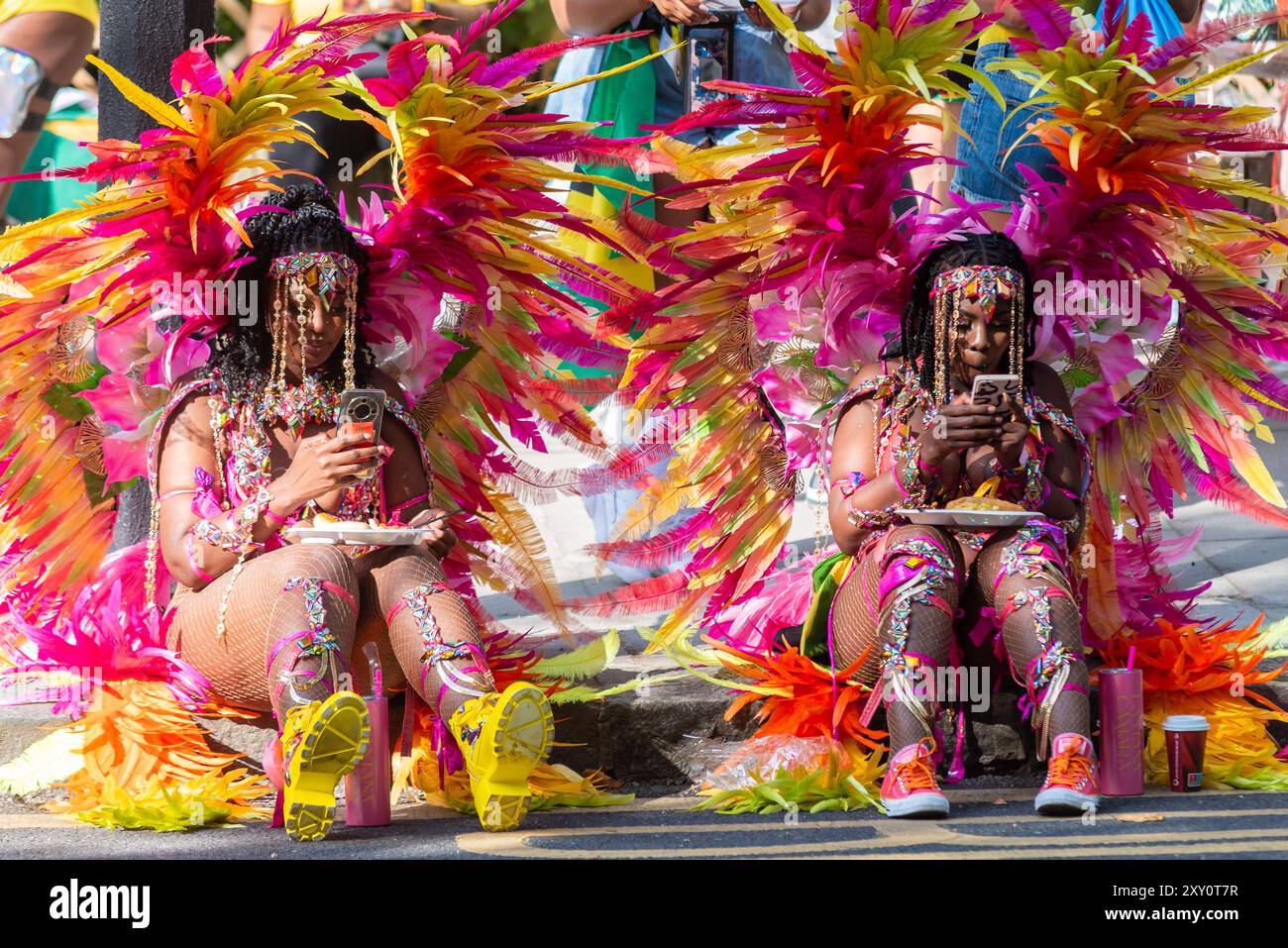 Zwei schwarze Frauen sitzen mit Essen und Telefon vor der Notting Hill Carnival Grand Parade 2024. Erwachsenentag am Montag an Feiertagen. Vorbereitungen Stockfoto