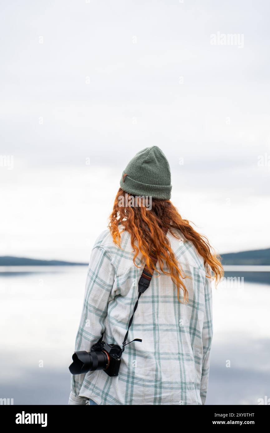 Eine junge Frau mit rotem Haar, die eine grüne Mütze und ein kariertes Hemd trägt, hält eine Kamera an einem ruhigen See. Ihr Rücken zeigt der Kamera, während sie die Erbse genießt Stockfoto