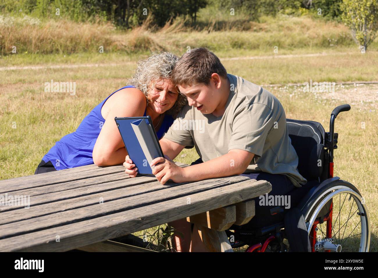 Ein kleiner Junge mit Zerebralparese, sitzt im Rollstuhl und verbindet sich draußen über ein digitales Tablet mit seiner Mutter Stockfoto