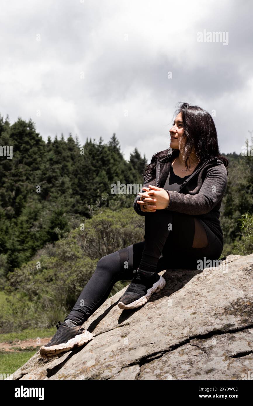 Eine Frau entspannt sich auf einem Felsen und blickt von der Kamera weg, umgeben von einer üppigen Waldkulisse. Sie wirkt zufrieden und nachdenklich, während sie den einnimmt Stockfoto