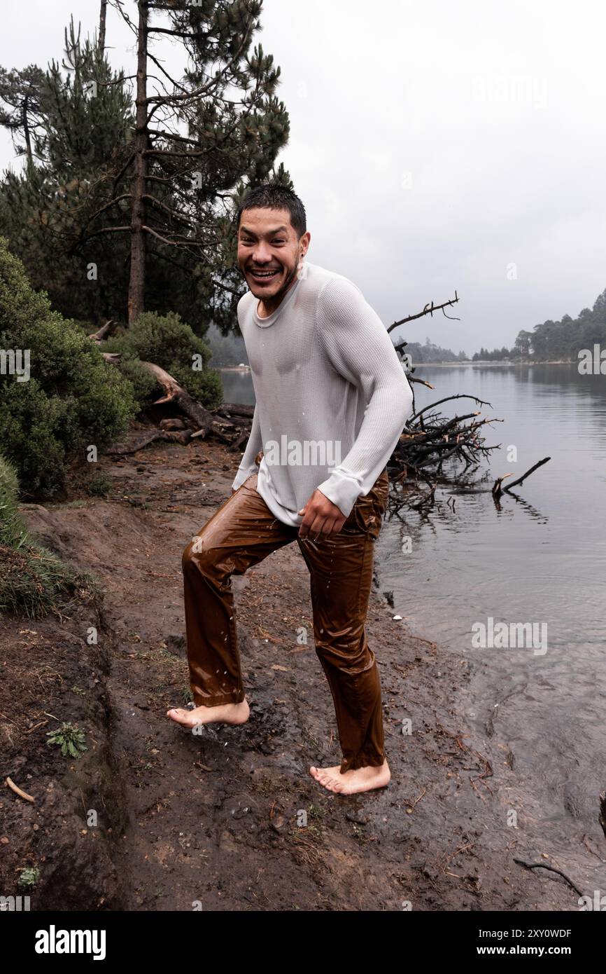 Ein fröhlicher lateinamerikanischer Mann, der an einem nassen und regnerischen Tag aus einem nassen See steigt und lächelt, umgeben von üppigen Kiefern und Nebel Stockfoto