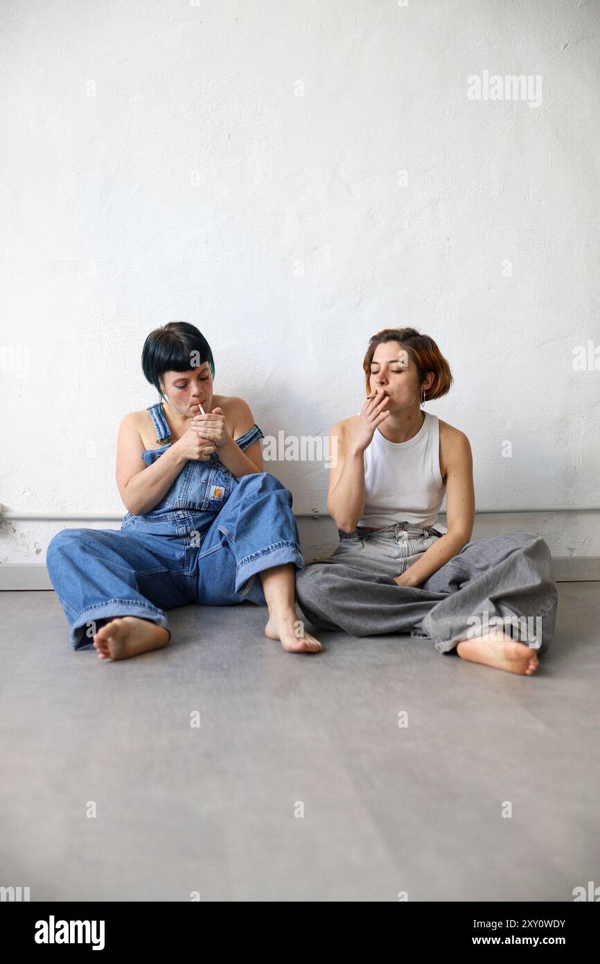 Zwei erwachsene Schwestern sitzen auf einem grauen Boden vor einer weißen Wand und rauchen eine Zigarette. Beide sind in entspanntem Denim gekleidet Stockfoto