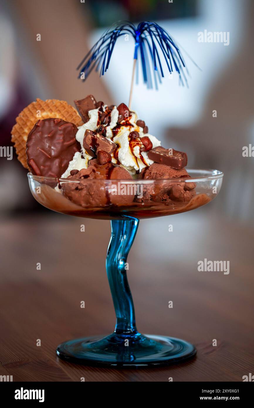 Ein luxuriöser Schokoladeneis-Eisbecher mit Schlagsahne, Nüssen und Keksen mit Schokoladenüberzug, garniert mit einem festlichen blauen Partyschirm. Stockfoto