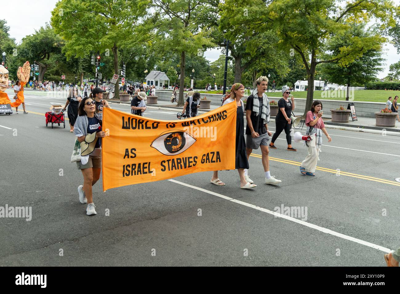 Washington DC, USA - 19.08.2024: Eine Gruppe von Menschen läuft die Straße hinunter und hält ein großes orangefarbenes Schild mit der Aufschrift: „Die Welt beobachtet, wie Israel hungert Stockfoto