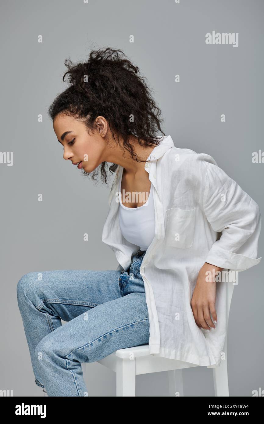 Eine junge Frau mit lockigen Haaren sitzt auf einem weißen Hocker, trägt ein weißes Hemd und blaue Jeans. Ihr Blick richtet sich nachdenklich nach unten. Stockfoto