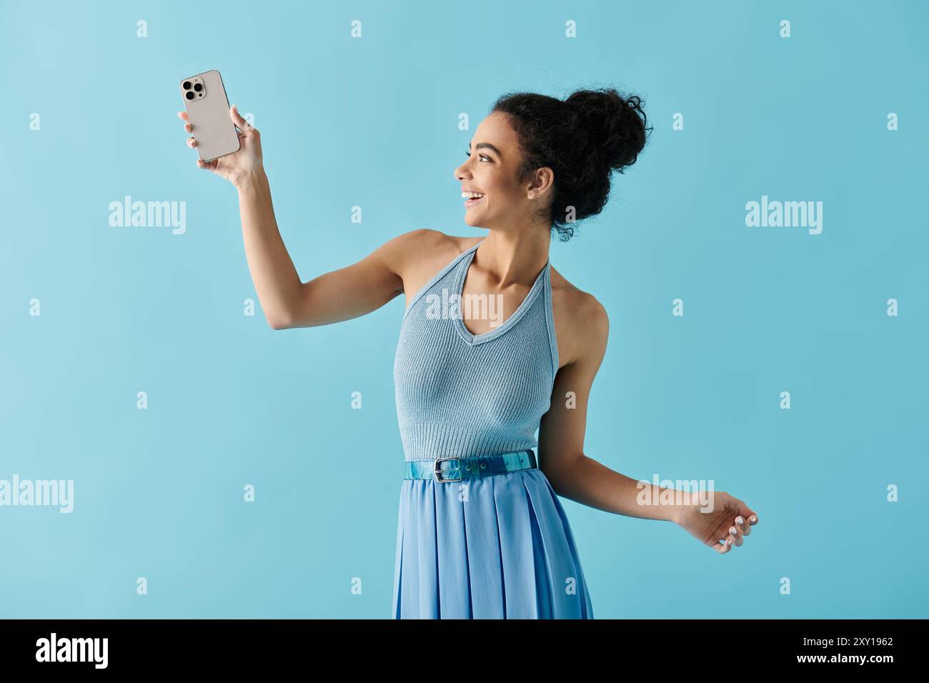 Eine junge Afroamerikanerin in einem blauen Kleid posiert für ein Selfie vor hellblauem Hintergrund. Stockfoto