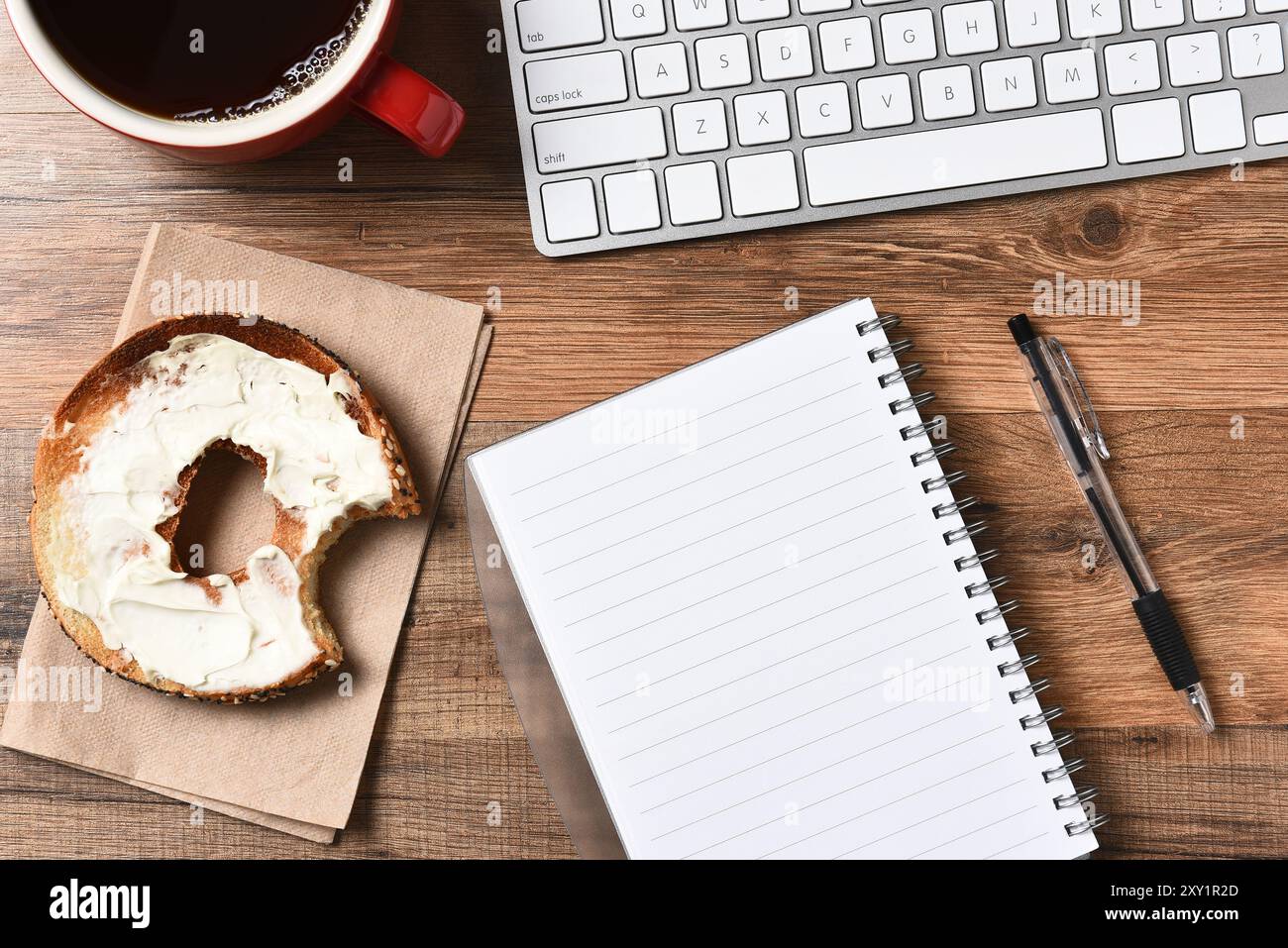 Ein Bagel mit Frischkäse und einem Biss, der neben einer Computertastatur und einem Pad und Stift herausgenommen wurde. Eine Tasse Kaffee in der oberen Ecke, Horizontal für einen Hi Stockfoto