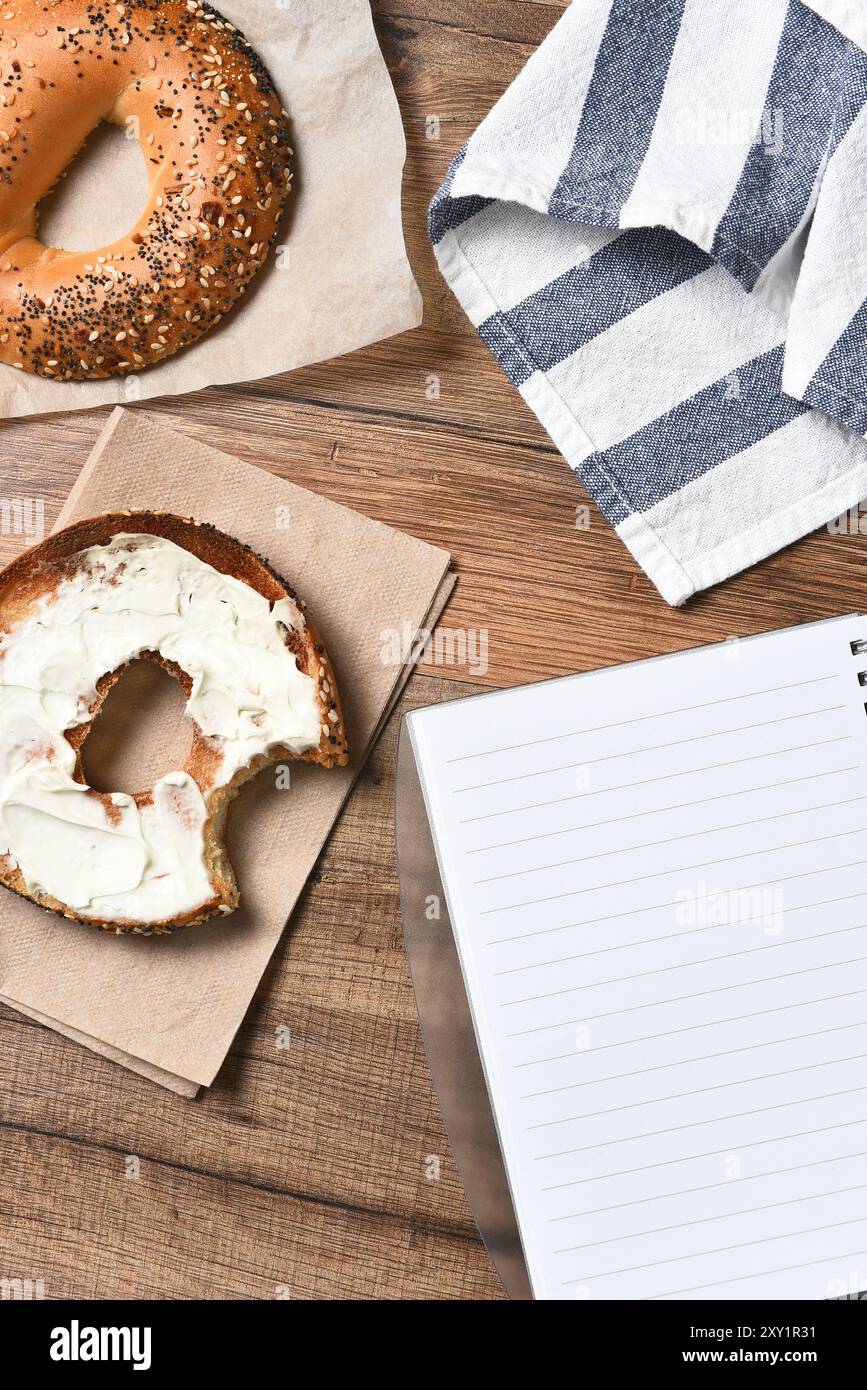 Ein Bagel mit Frischkäse und einem Biss. Mit Handtuch und Notizblock auf einem rustikalen Holzschreibtisch. Hochwinkelansicht im vertikalen Format, Stockfoto