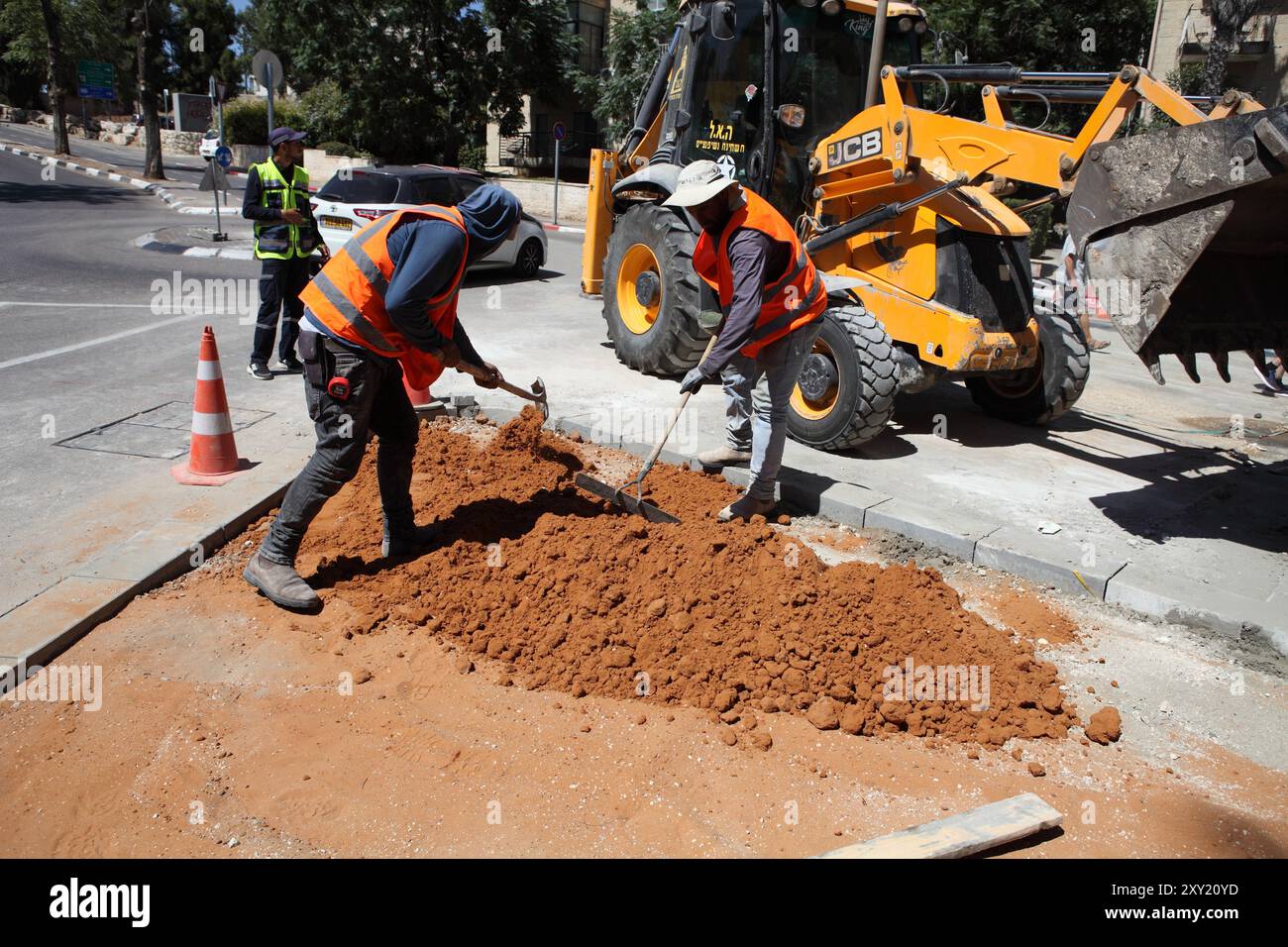 Die Arbeiter tragen Schutzwesten, einer benutzt einen Siebkasten, der andere einen Spaten, sie verteilen roten Boden auf dem Bürgersteig, damit Steine darauf gelegt werden können, wenn der Bürgersteig wieder aufgebaut wird Stockfoto