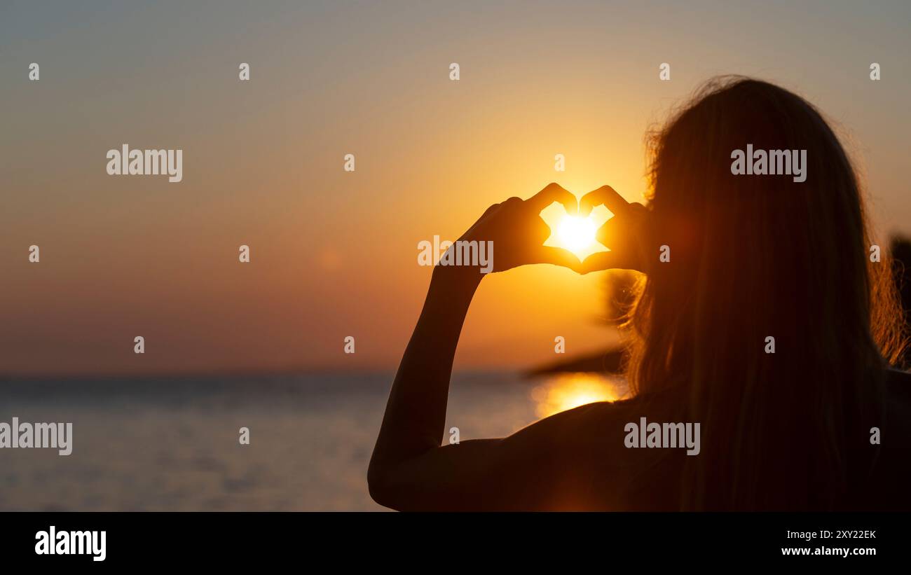 Blick über die Schulter einer jungen Frau, die mit ihren Fingern eine Herzform mit der untergehenden oder aufgehenden warmen orangefarbenen Sonne, die durch das hindurch scheint, bildet Stockfoto