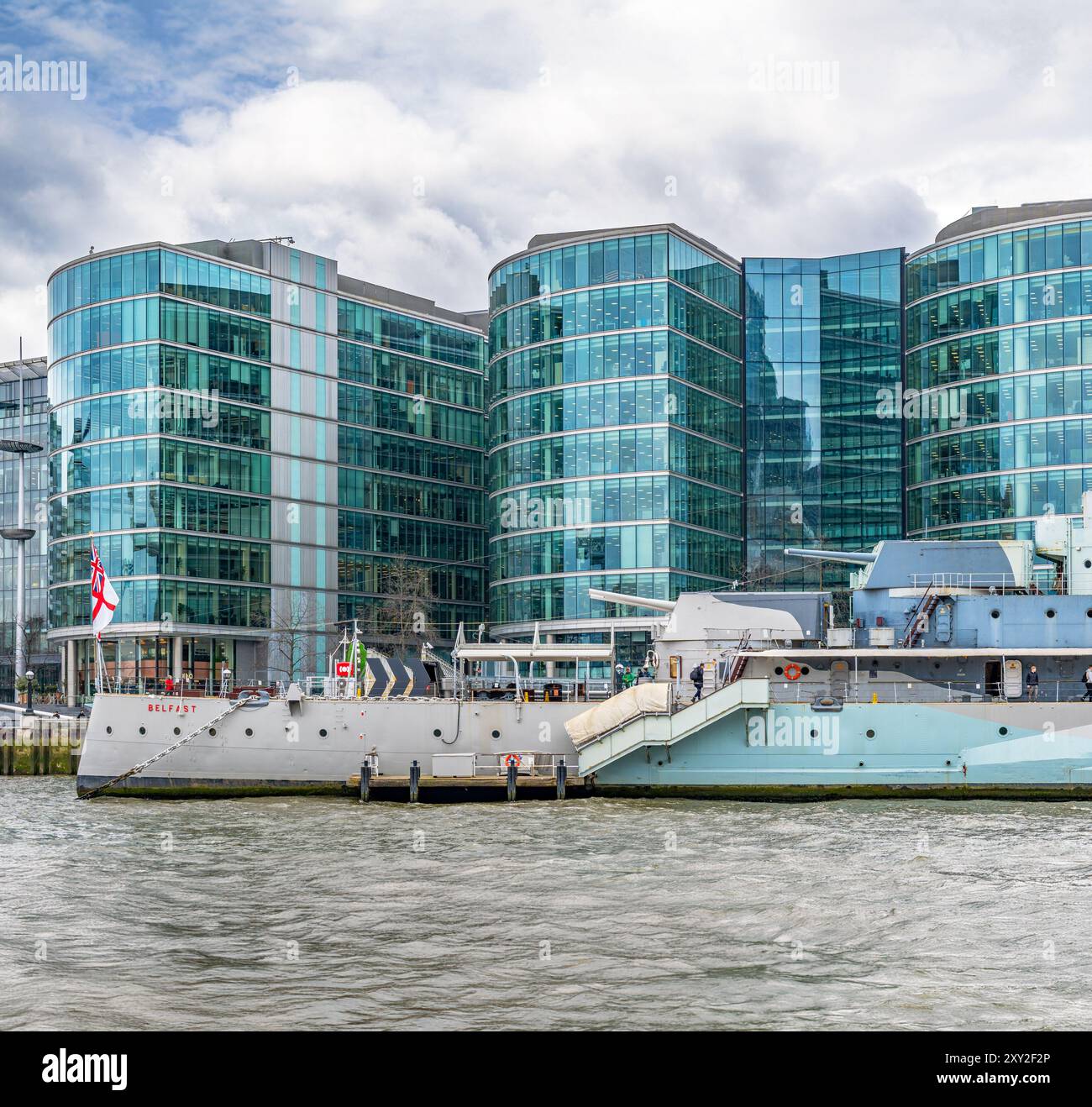 Seitenansicht des schwimmenden Kriegsmuseums HMS Belfast, das an der Themse angedockt ist, mit Touristen, die die Außendecks unter einem regnerisch bewölkten Himmel flanieren. Stockfoto