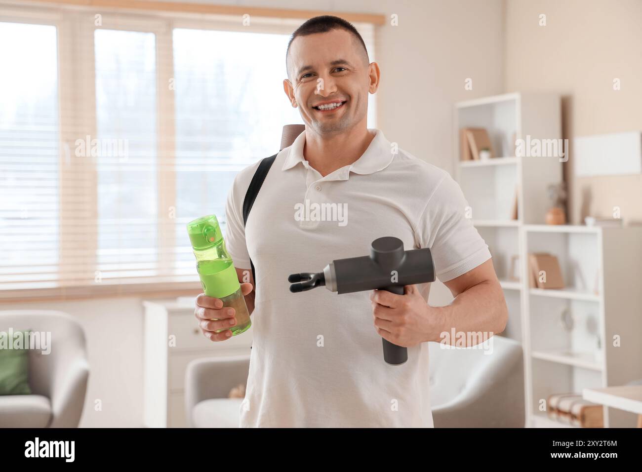 Sportlicher junger Mann mit perkussivem Massagegerät und Wasserflasche zu Hause Stockfoto