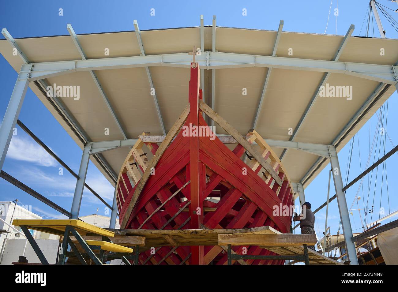 Vorderansicht eines teilweise gebauten roten Holzschiffes in einer überdachten Werft unter klarem Himmel, Patmos Marine, Bootswerft, Diakofti, Patmos, Dodekanesisch, Griechisch Stockfoto