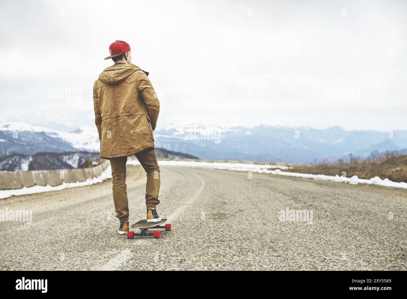 Stylischer Happy Young man in einer Mütze und Hose Jogginghose, die auf einem Longboard eine Bergstraße hinunterrollt und das Leben genießt Stockfoto