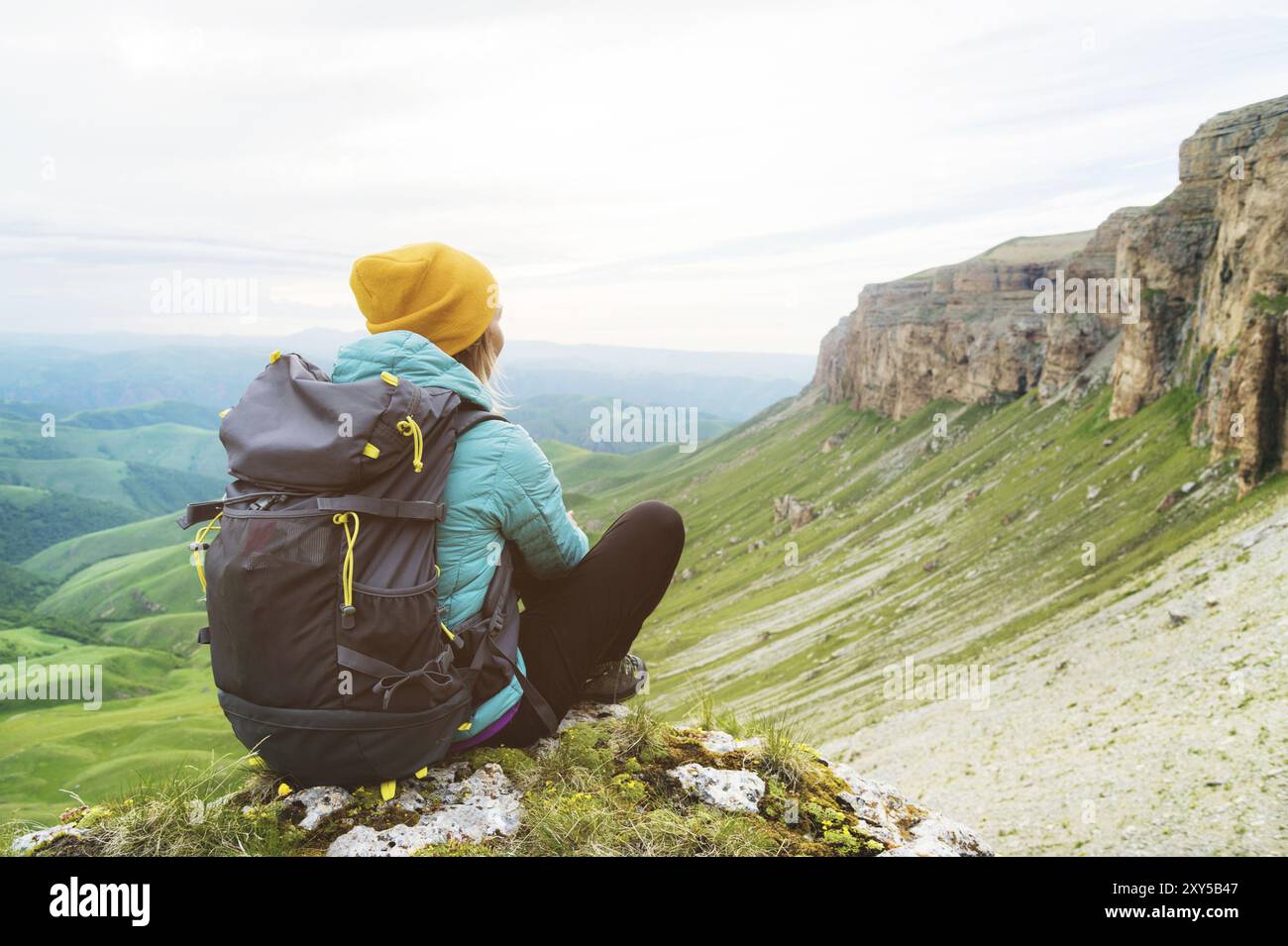 Zurücksitzendes Mädchen mit gelbem Hut und einer Sonnenbrille, die am Fuße epischer Felsen mit einem Rucksack sitzt und wegschaut Stockfoto