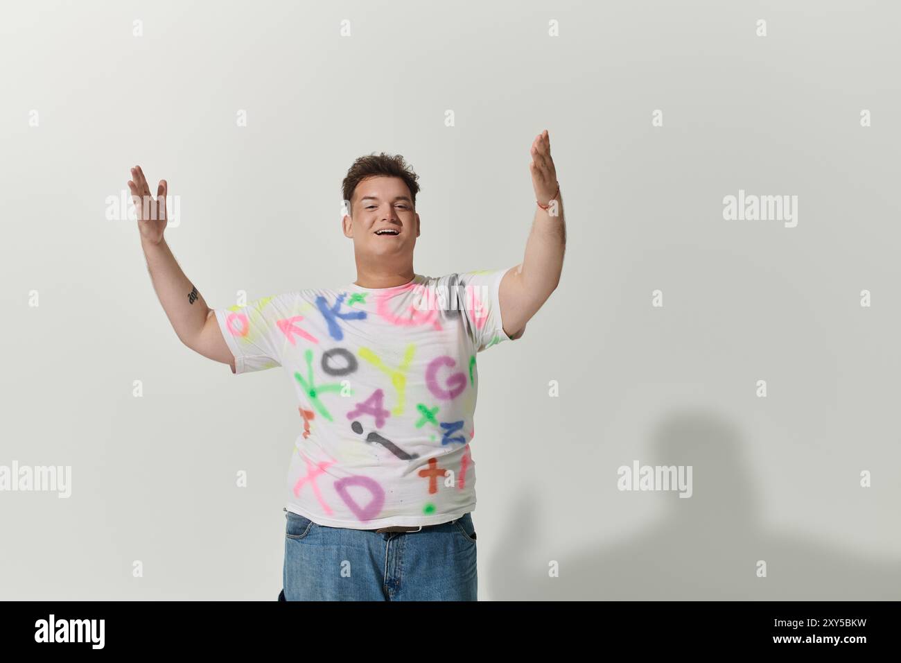 Queere Person mit kurzen braunen Haaren, großem Lächeln, weißem T-Shirt mit bunten Buchstaben, Arme vor Freude erhoben Stockfoto