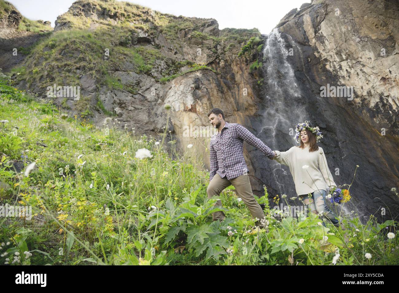 Hipster mit Bart führt seine Braut in die Natur vor dem Hintergrund eines Wasserfalls, der von einem Felsen fällt Stockfoto