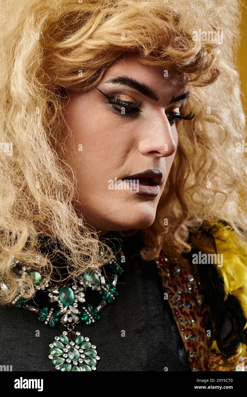 Blonde queere Person in dramatischem Make-up schaut nach unten, trägt smaragdgrüne Halskette Stockfoto