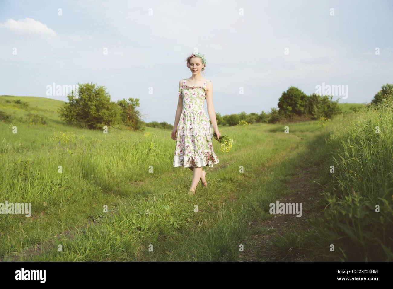 Ein junges glückliches lächelndes Mädchen in einem Calico-Kleid mit einem Blumenstrauß läuft auf einer Landstraße mit grünem Gras Stockfoto