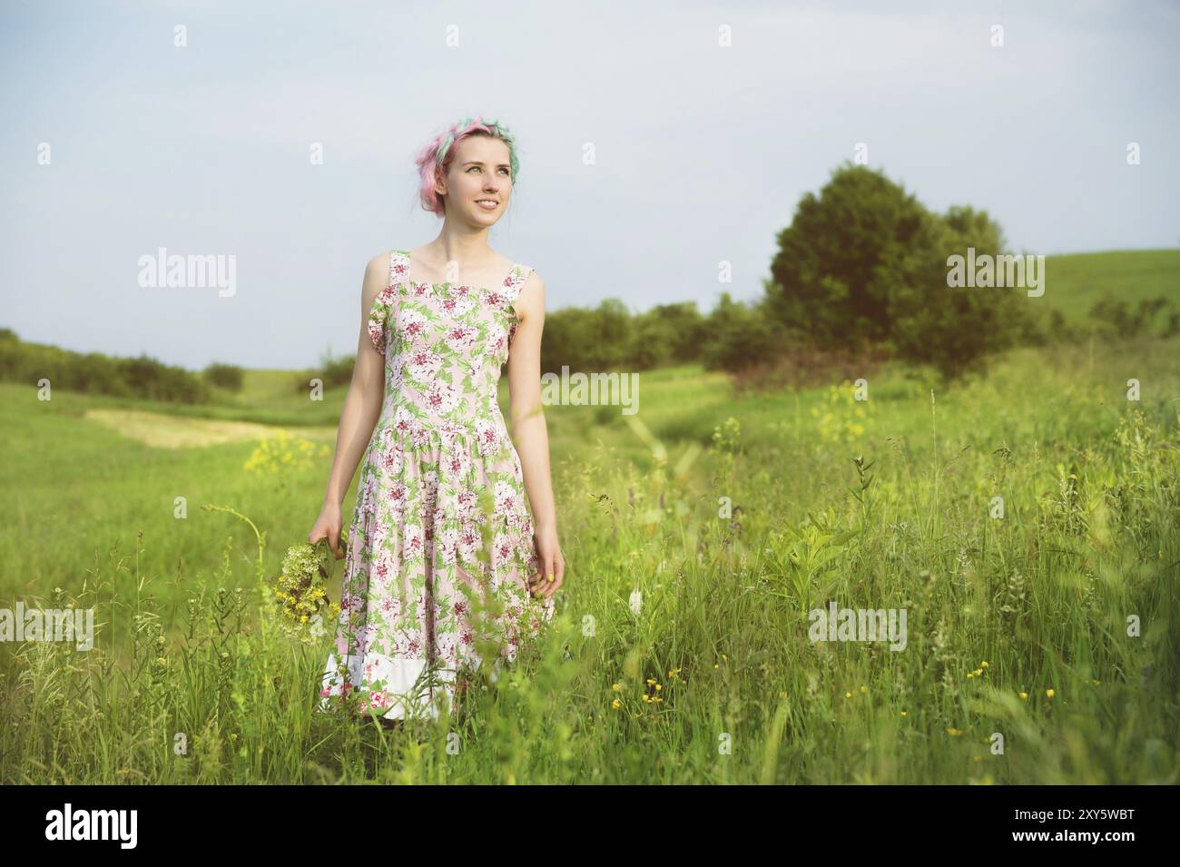 Ein junges glückliches lächelndes Mädchen in einem Calico-Kleid mit einem Blumenstrauß läuft auf einer Landstraße mit grünem Gras Stockfoto