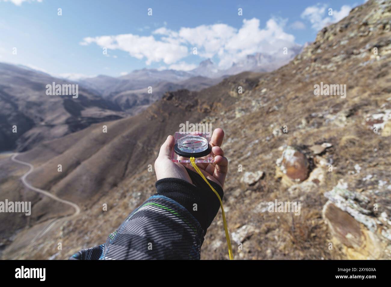 Die Hand eines Mannes hält einen magnetischen Taschenkompass für die Navigation vor einem felsigen Hang und epischen Felsen unter einem blauen Himmel und weißen Wolken. Stockfoto