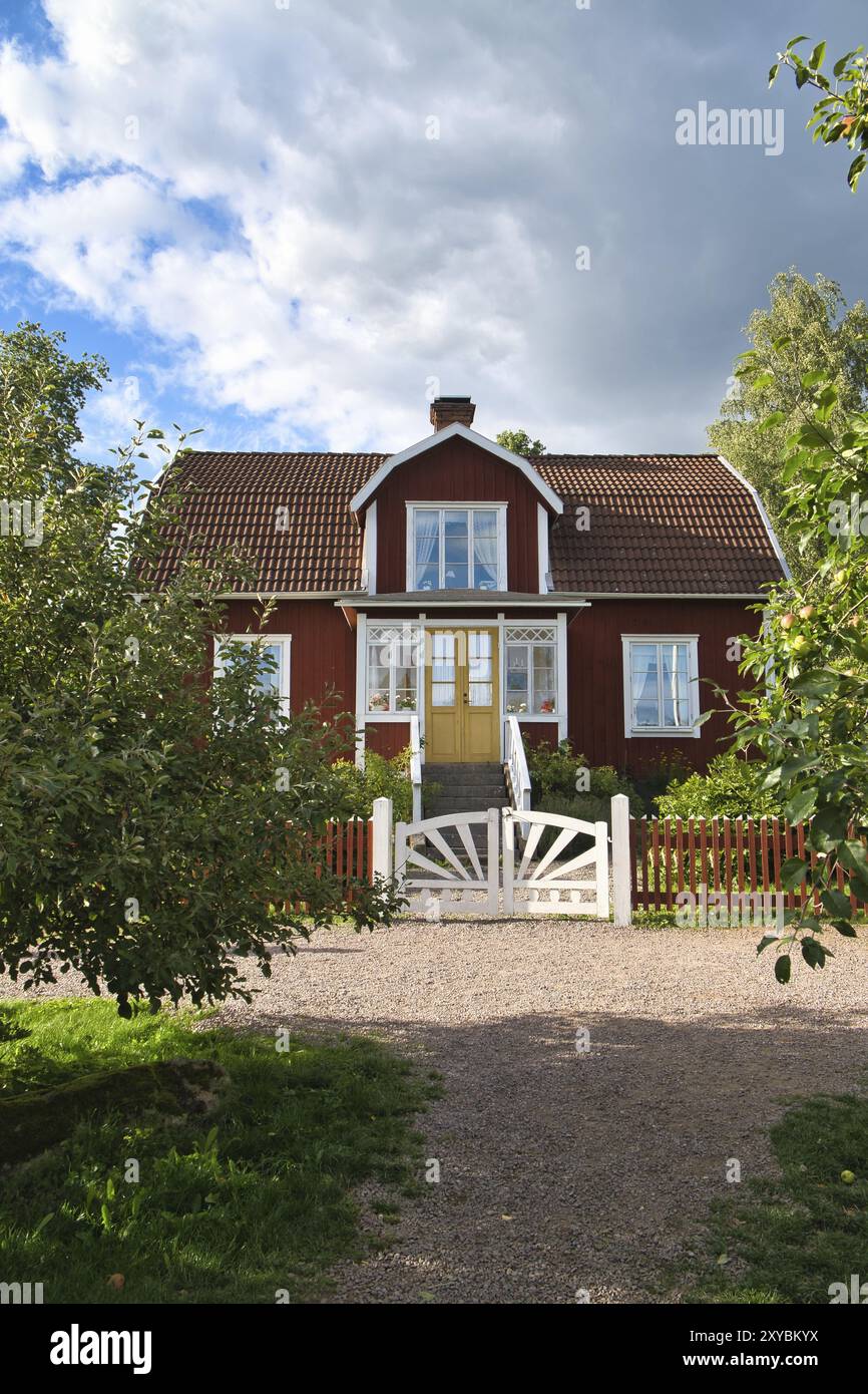 Ein typisches rot-weißes schwedisches Haus im kleinen und kleinen Stil. Weißes Gartentor, brauner Zaun. Grüner Rasen im Garten. Bäume im Vordergrund. Blauer Himmel mit sm Stockfoto