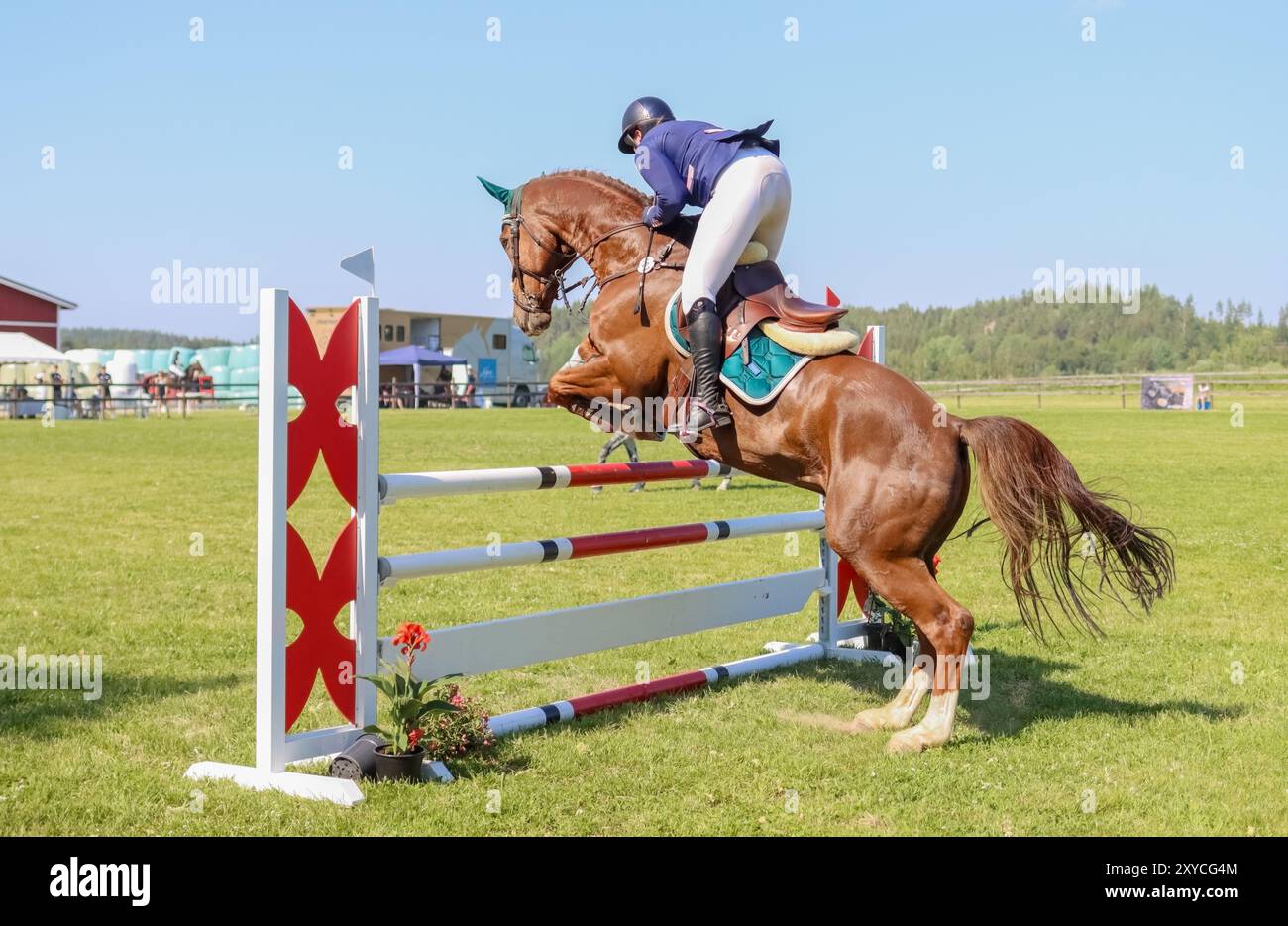 Reitwettbewerbe in Lohja, Finnland. Ein Fahrer überquert an einem sonnigen Tag ein Hindernis. Im Hintergrund befindet sich ein Publikum. Eine Frau reitet auf einem Pferd. Stockfoto