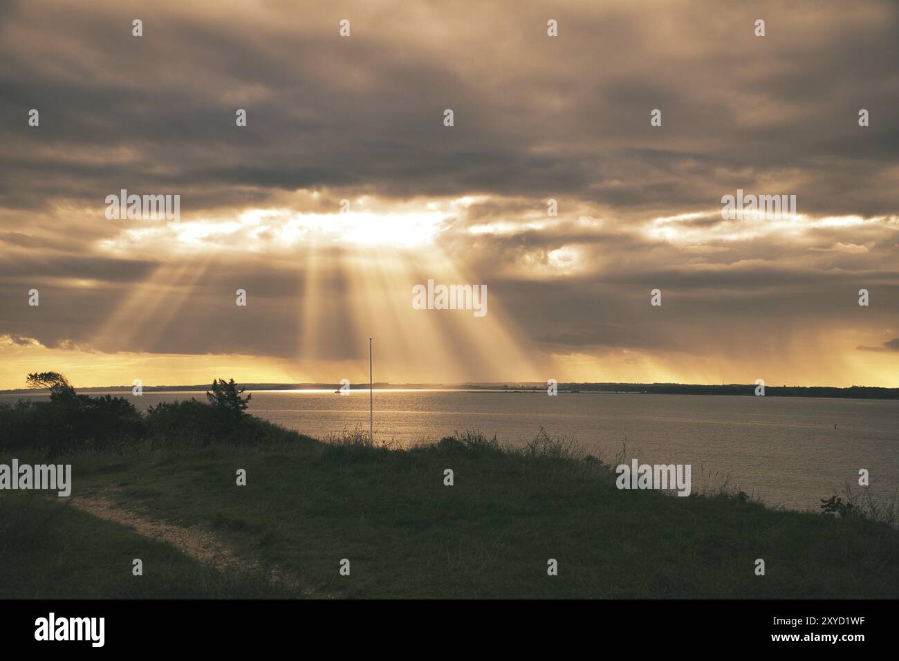 An der Küste von Hundested. Sonnenstrahlen durchbrechen den dramatischen Himmel durch die Wolken. Wiese mit Pfad im Vordergrund. Landschaftsaufnahmen in Dänemark Stockfoto