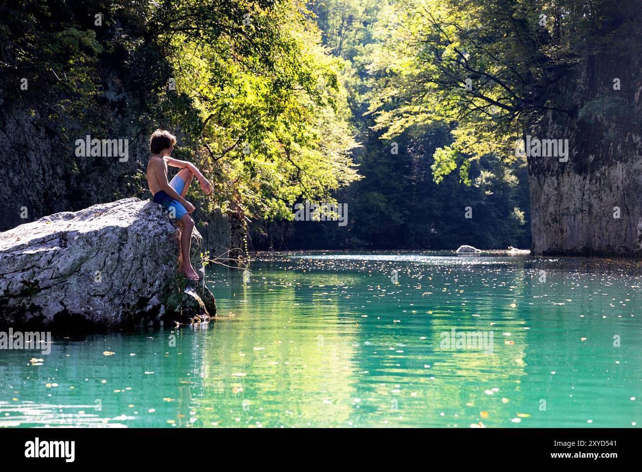 Ein Junge in Badehose, der auf einem Felsen mitten im Idrijca-Fluss-Canyon in der Nähe von Most na Soči, Slowenien, an einem sonnigen heißen Sommertag sitzt Stockfoto