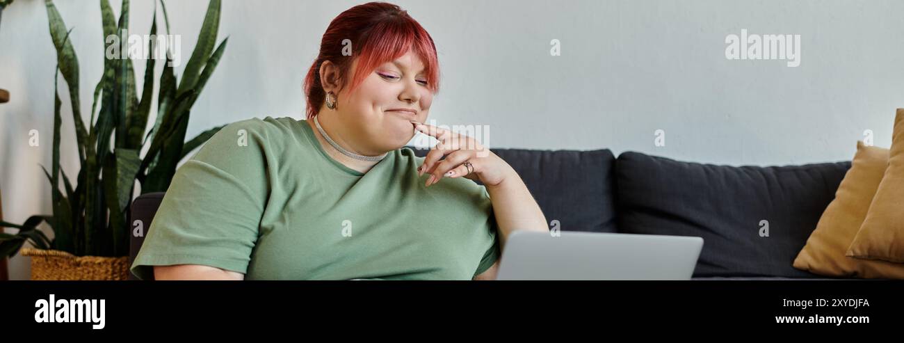 Eine große Frau mit rosa Haaren sitzt auf einer Couch in ihrem Haus und lächelt, während sie auf einen Laptop schaut. Stockfoto