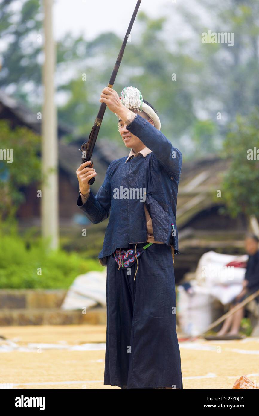 Biasha, China, 17. September 2007: Miao ethnische Minderheit in traditioneller blauer Kleidung, geflochtenem Kopfschmuck und langem Haarbutt, der eine Waffe in die Luft schießt Stockfoto