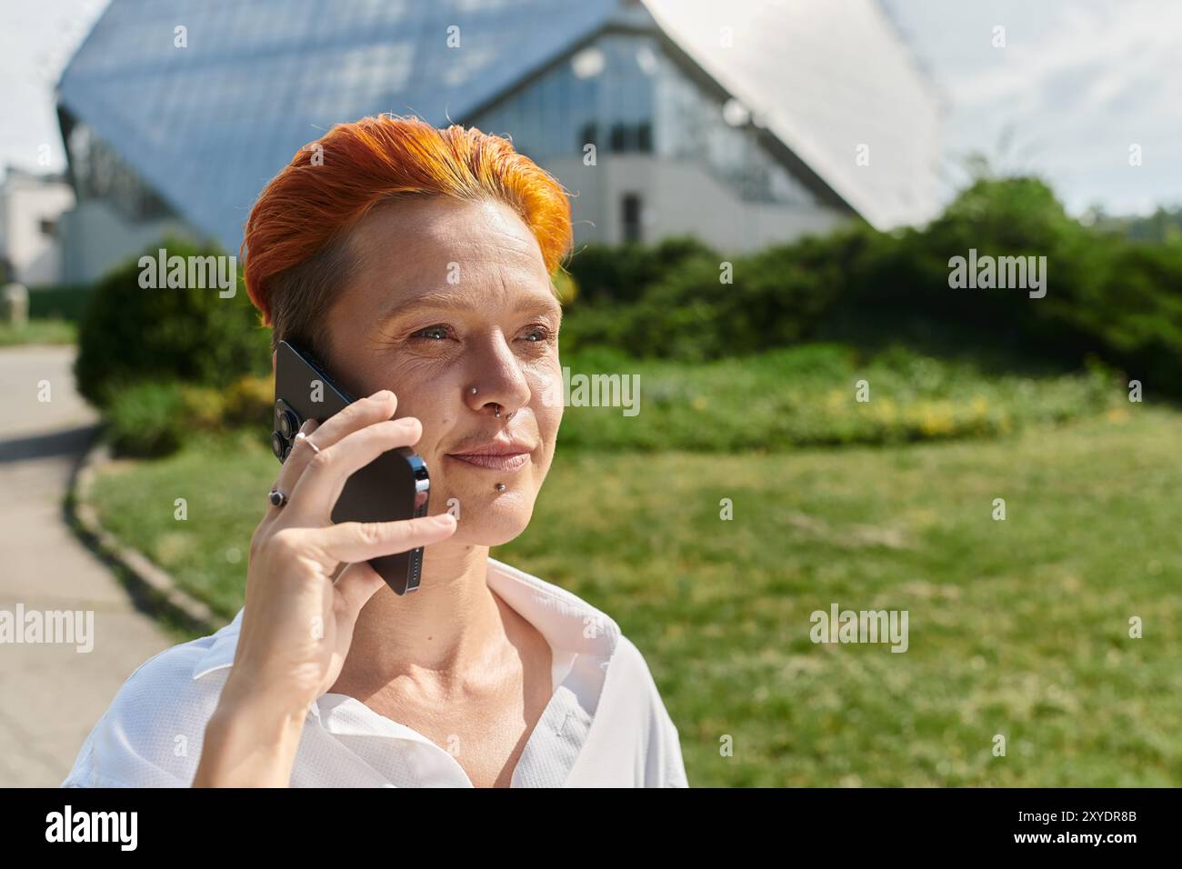 Eine Frau mit orangefarbenen Haaren redet auf ihrem Telefon, während sie auf einem sonnigen College-Campus läuft. Stockfoto