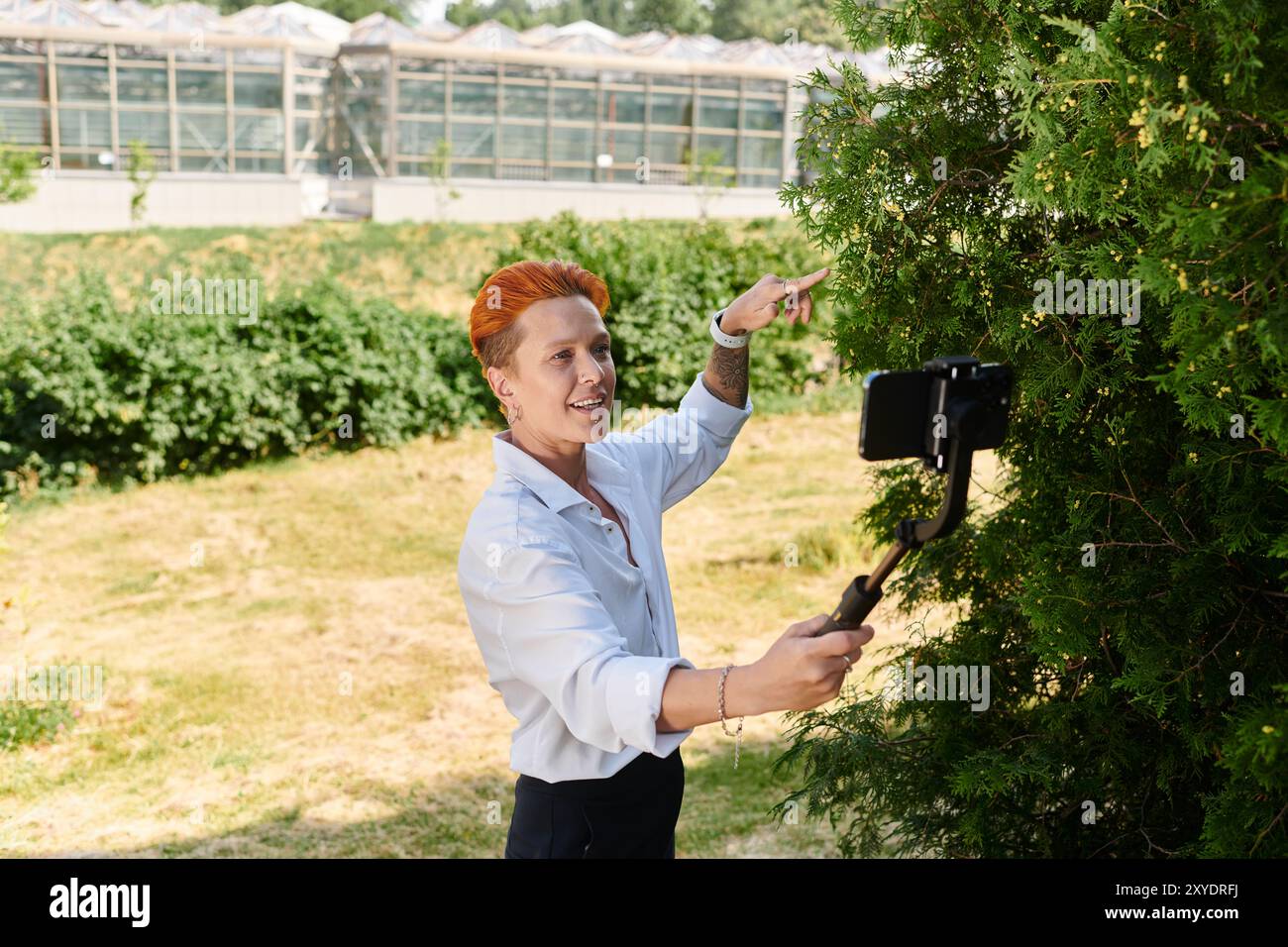 Frau mit orangefarbenen Haaren filmt sich am Telefon um Baum auf dem Campus Stockfoto