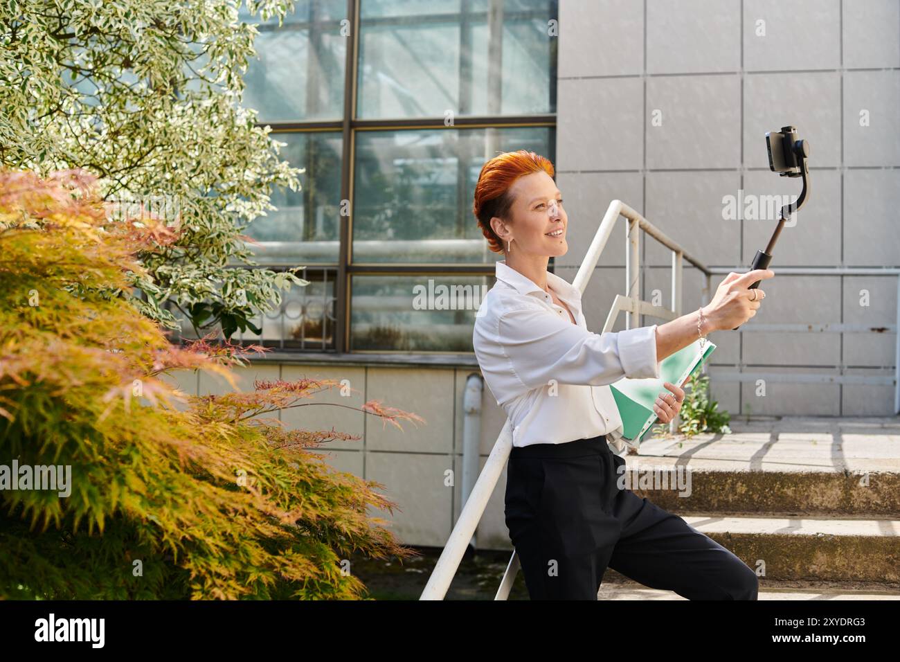 Eine Frau lächelt und hält ein Handy auf einem Selfie-Stick, lehnt sich an einem sonnigen Tag an ein Geländer. Stockfoto