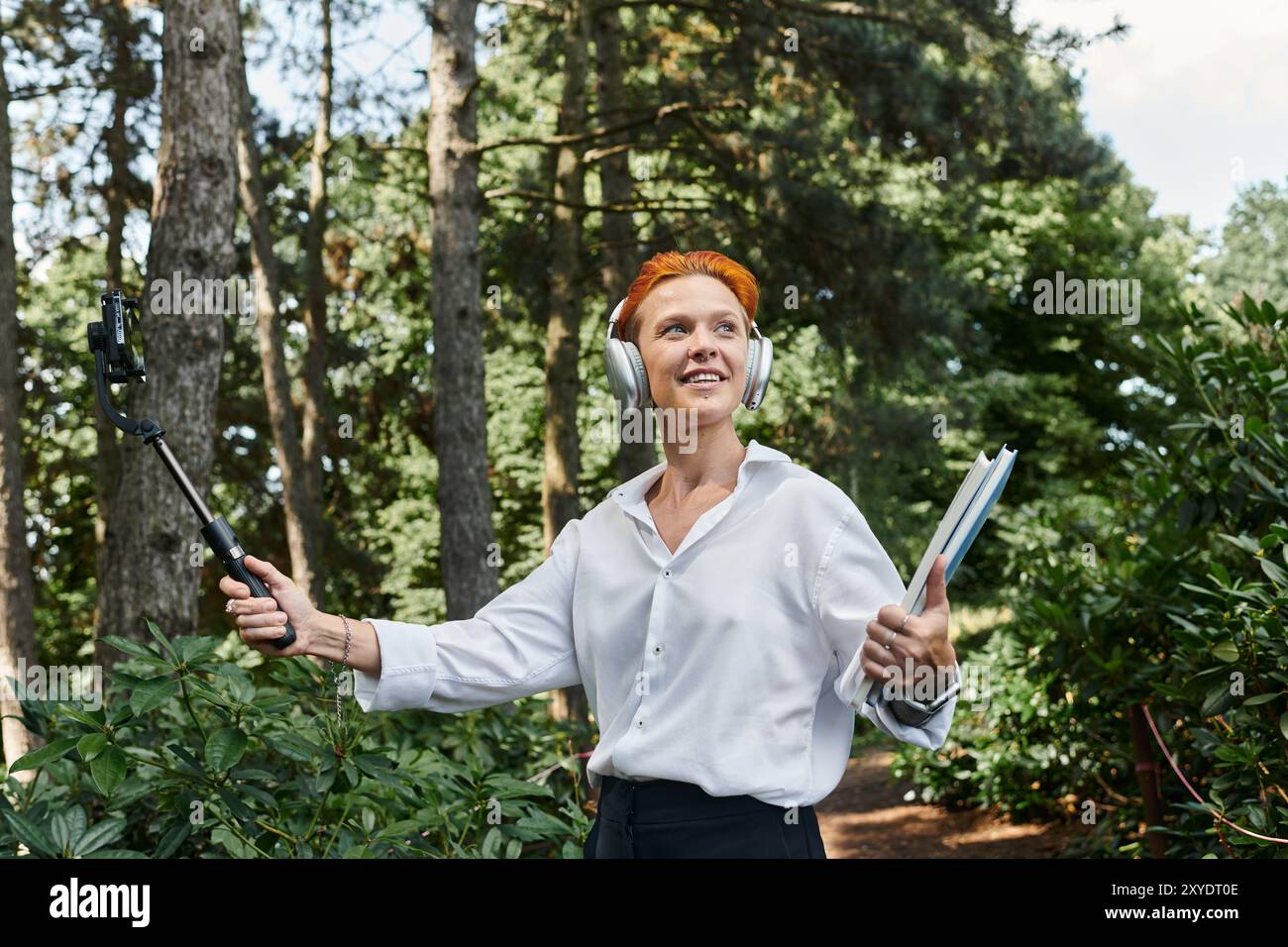 Ein Lehrer in weißem Hemd und Kopfhörern geht durch einen Campus, hält einen Selfie-Stick und Bücher. Stockfoto